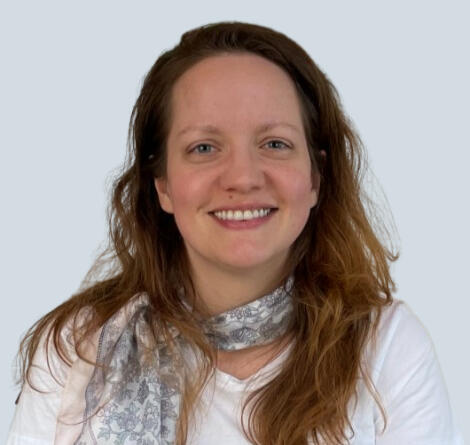 A smiling woman with long light-brown hair, wearing a white shirt and a patterned scarf, posed against a light blue background.